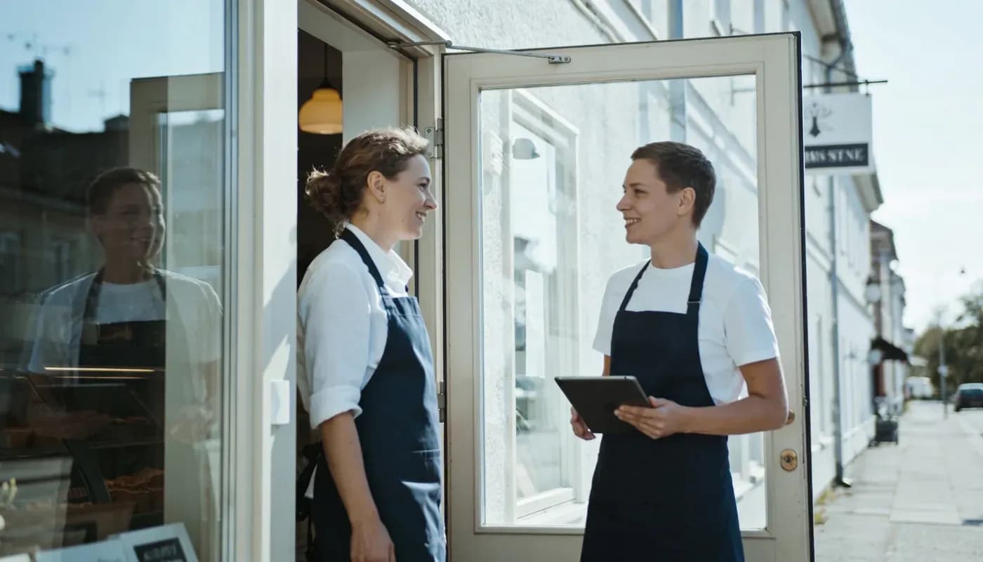 Kaksi yrittäjää keskustelee liikkeen ovella aamuvalossa, toisella tabletti kädessään.